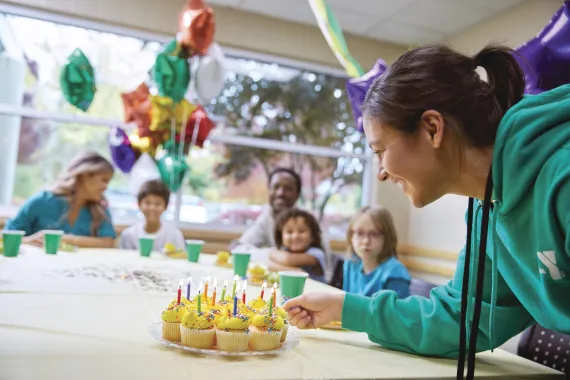 a group of children surrounded by balloons and a Y employee lighting the candles on a platter of cupcakes.