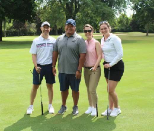 2 men and 2 women smiling and holding golf clubs while standing on the putting green