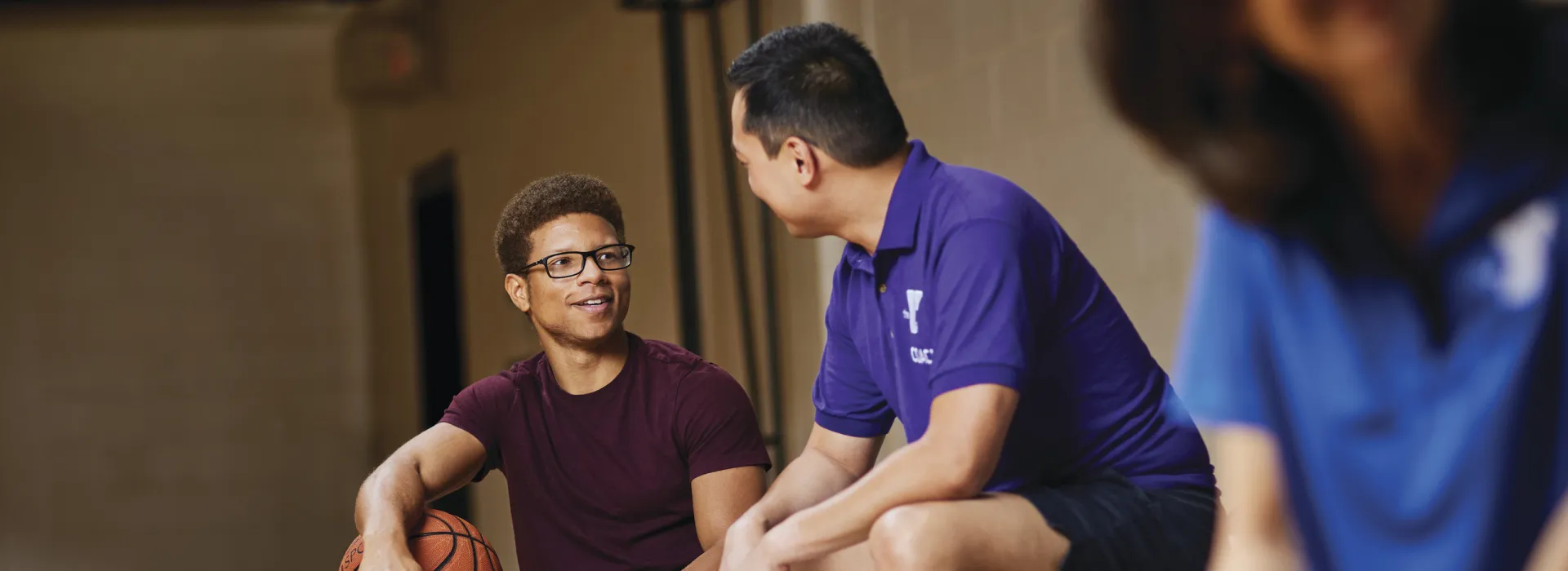 A young man in a YMCA staff shirt sitting on the bleachers smiling and talking with another young man who is there to play basketball.