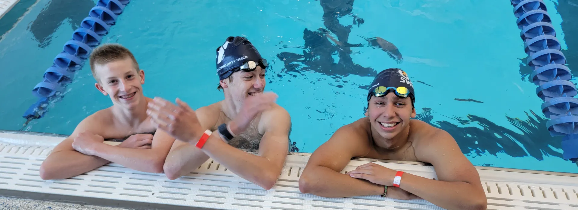 3 young boys in swim caps treading water pool side, smiling and enjoying the pool.