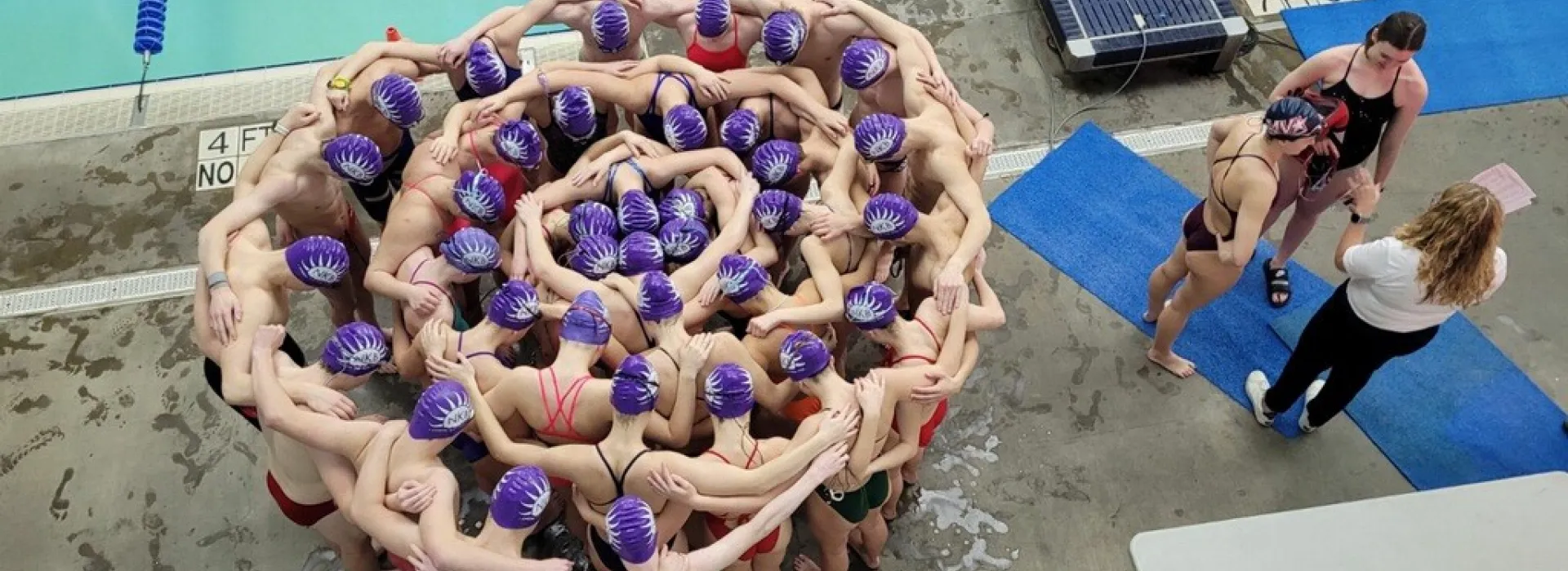 more than 30 swimmers wearing swim caps forming a circle on the pool deck with their hands around each other.