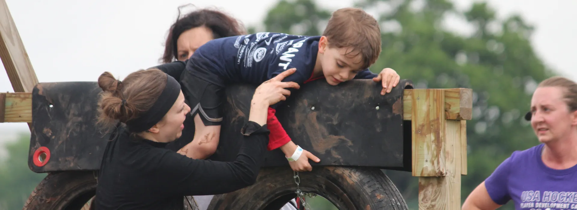 A mud run volunteer helping a young boy over an obstacle.